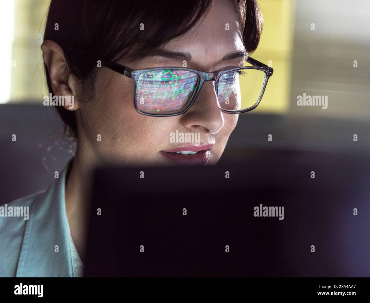 Eine fokussierte Person, die eine Brille mit Computercode-Reflexion in den Objektiven trägt, schaut intensiv auf einen Computerbildschirm. Stockfoto