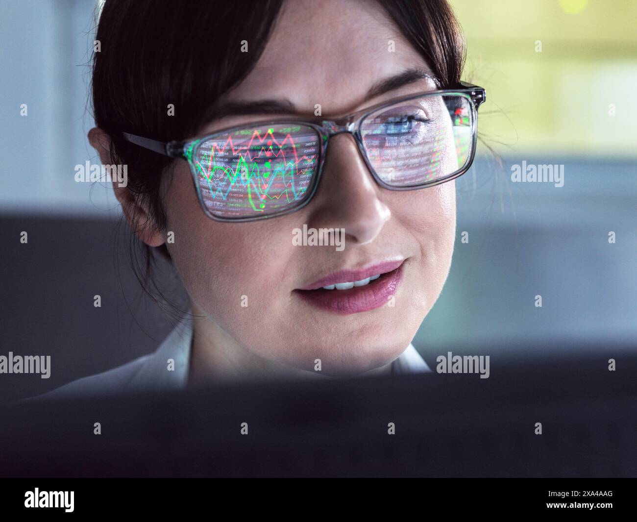 Eine Frau mit einer Brille, die bunte Datengrafiken reflektiert, schaut auf einen Computerbildschirm mit einem fokussierten Ausdruck. Stockfoto