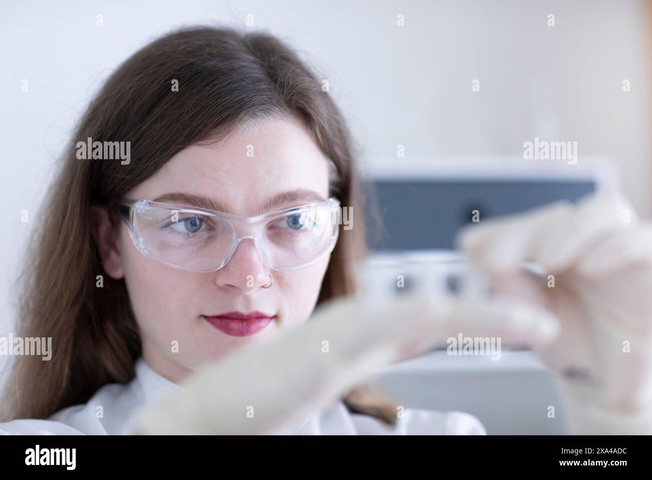Eine fokussierte Frau, die eine Schutzbrille trägt, untersucht ein Fläschchen im Labor. Stockfoto