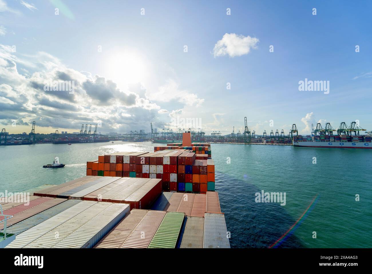 Ein Frachtschiff ist mit bunten Containern unter einem teilweise bewölkten Himmel beladen und fährt in der Nähe eines belebten Hafens mit zahlreichen Kränen und Schifffahrtsinfrastruktur. Stockfoto