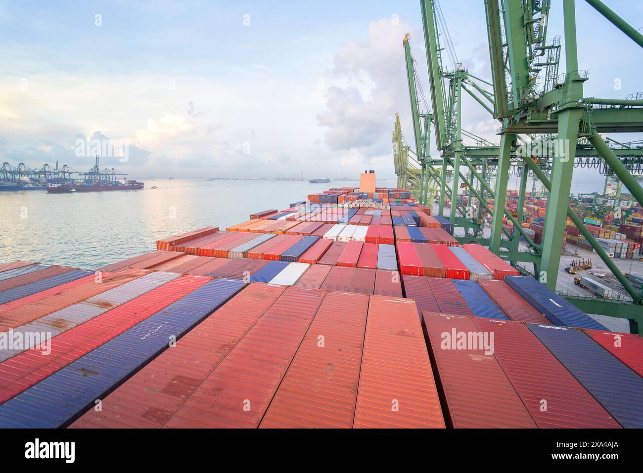 Ein Containerschiff wird an einem belebten Hafen mit klarem Himmel bei Sonnenaufgang oder Abenddämmerung angedockt, während Containerkräne über die bunte Vielfalt von Containern ragen. Stockfoto