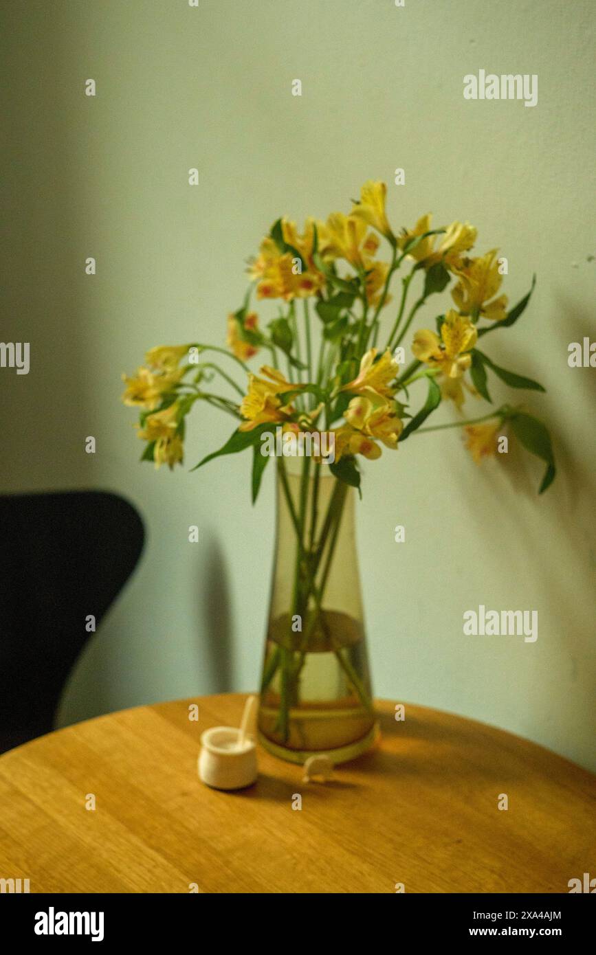 Eine Vase mit gelben Blumen sitzt auf einem Holztisch vor einer hellen Wand, begleitet von einem kleinen Holzgegenstand. Stockfoto
