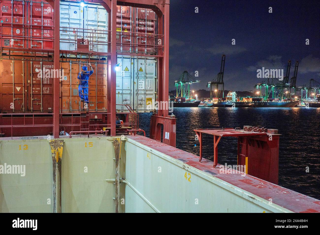 Ein Frachtschiff legt nachts an einem belebten Hafen an, wobei Container auf Deck gestapelt sind und Kräne im Hintergrund sichtbar sind. Stockfoto