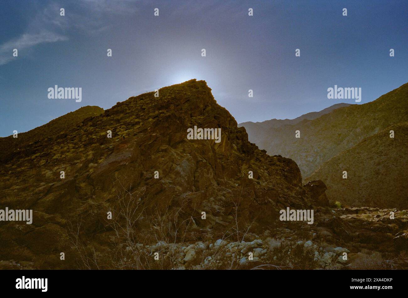 Die Sonne schwebt über dem Gipfel eines zerklüfteten Berges und strahlt ein warmes Licht auf die felsige Landschaft aus, während der klare Himmel in einen weichen Gefälle übergeht. Stockfoto