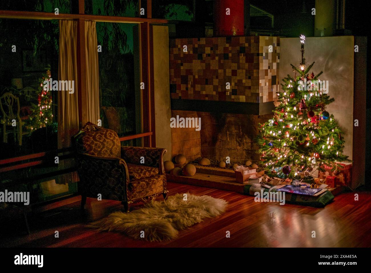 Ein gemütlicher Raum bei Nacht, beleuchtet vom warmen Leuchten eines festlichen Weihnachtsbaums mit bunten Lichtern und Ornamenten. Die Geschenke sind ordentlich angeordnet, neben einem dekorierten Kamin, mit einem bequemen Sessel an der Seite. Stockfoto