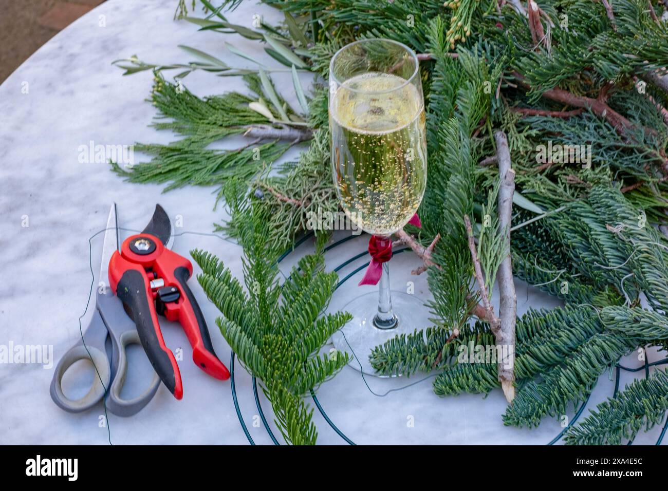 Ein Glas Sekt steht auf einer Marmorplatte neben einer Schere, Gartenschere und einer Reihe frischer Kiefernzweige, was ein Basteln oder Dekorieren im Urlaub andeutet. Stockfoto