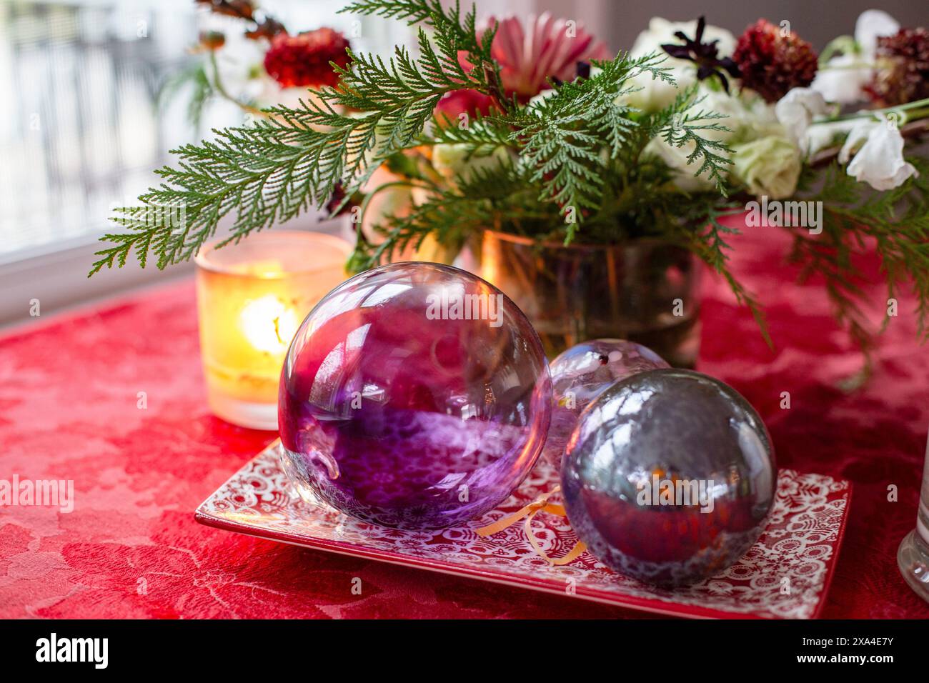 Eine festliche Dekoration mit glänzenden lila und silbernen Kugeln auf einer roten Tischdecke, begleitet von einer beleuchteten Kerze und einem Blumenstrauß, schafft eine warme und einladende Atmosphäre. Stockfoto