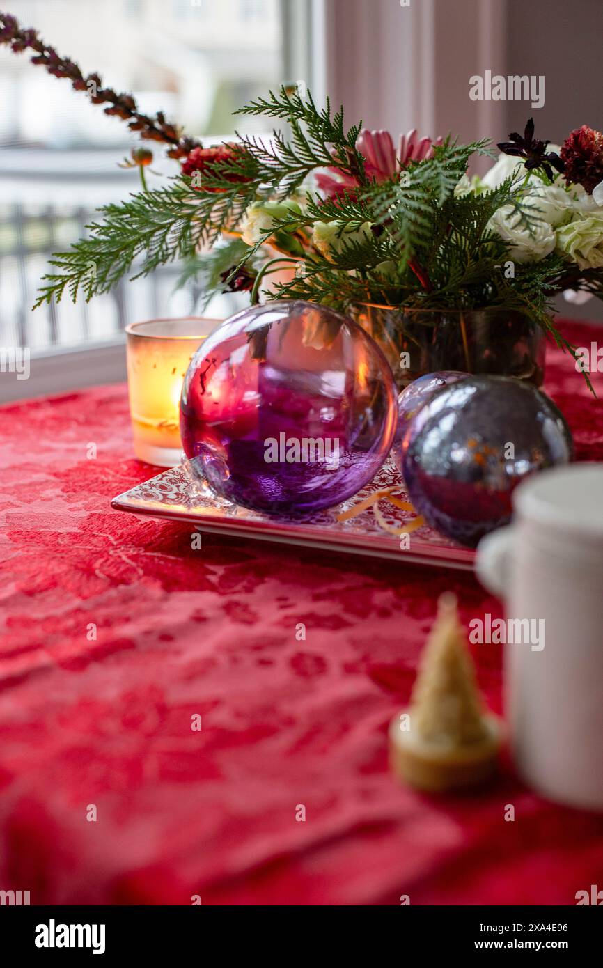 Ein festlicher Tisch mit einer leuchtend roten Tischdecke, die ein dekoratives Arrangement aus Blumen, Kerzen und bunten Kugeln auf einem silbernen Tablett neben einer weißen Kanne und einem kleinen dekorativen Baum zeigt. Stockfoto