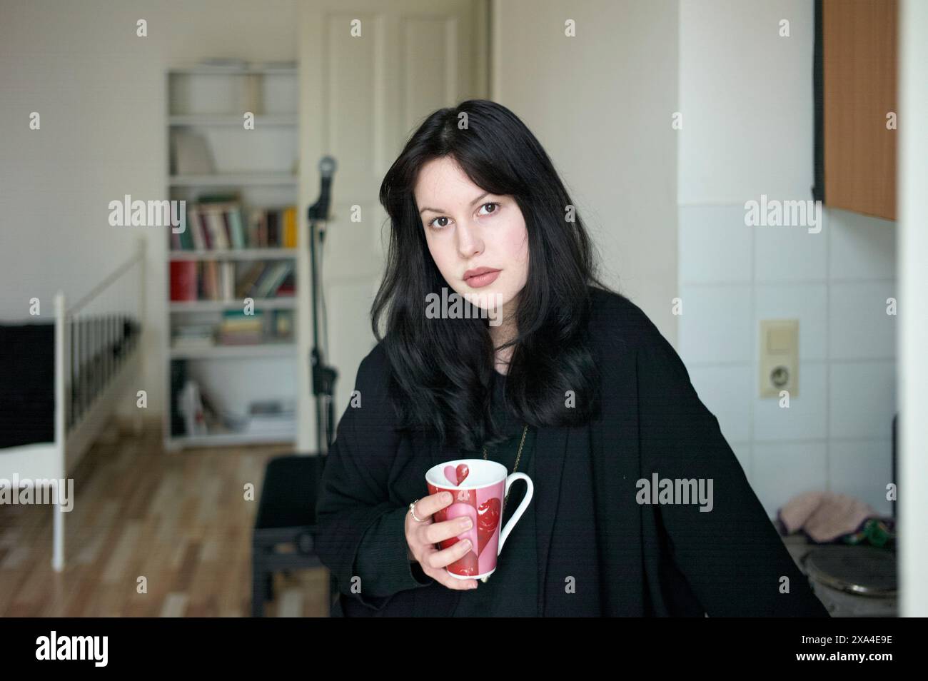 Eine junge Frau mit dunklem Haar steht drinnen mit einem roten Becher, trägt einen schwarzen Mantel mit einem neutralen Gesichtsausdruck. Stockfoto