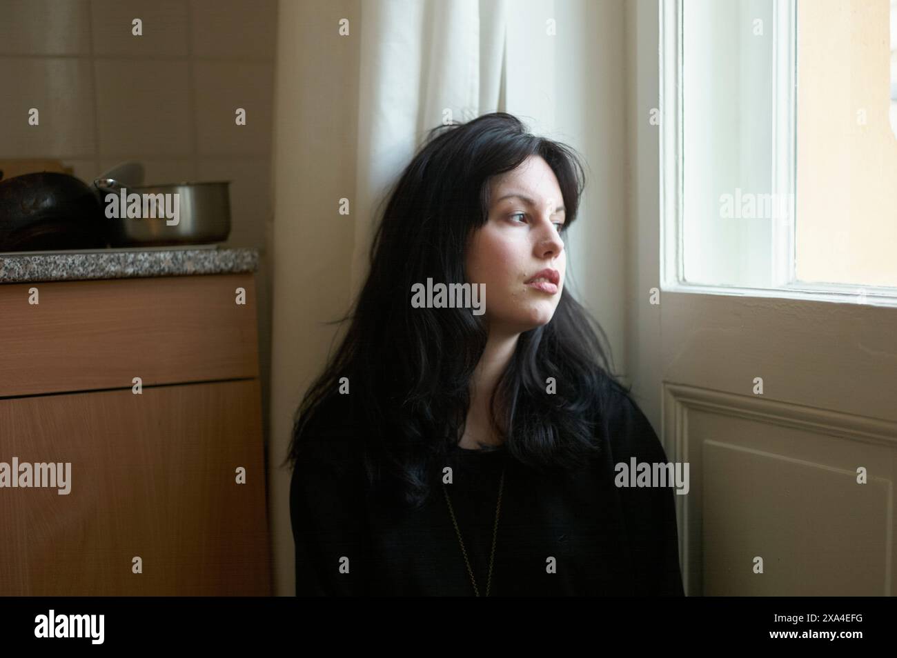 Eine Frau mit dunklen Haaren blickt aus einem Fenster in einem Raum, beleuchtet von natürlichem Licht. Stockfoto