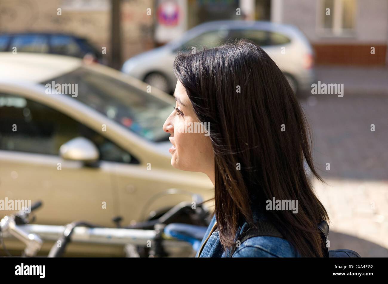 Eine Frau mit langen dunklen Haaren wird im Profil fotografiert, die an einem sonnigen Tag nach vorne blickt, mit Autos und Fahrrädern im Hintergrund. Stockfoto