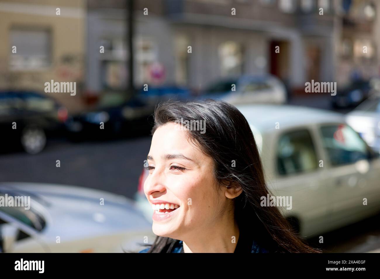 Eine lächelnde Frau draußen an einem sonnigen Tag, mit verschwommenen Straßen und geparkten Autos im Hintergrund. Stockfoto