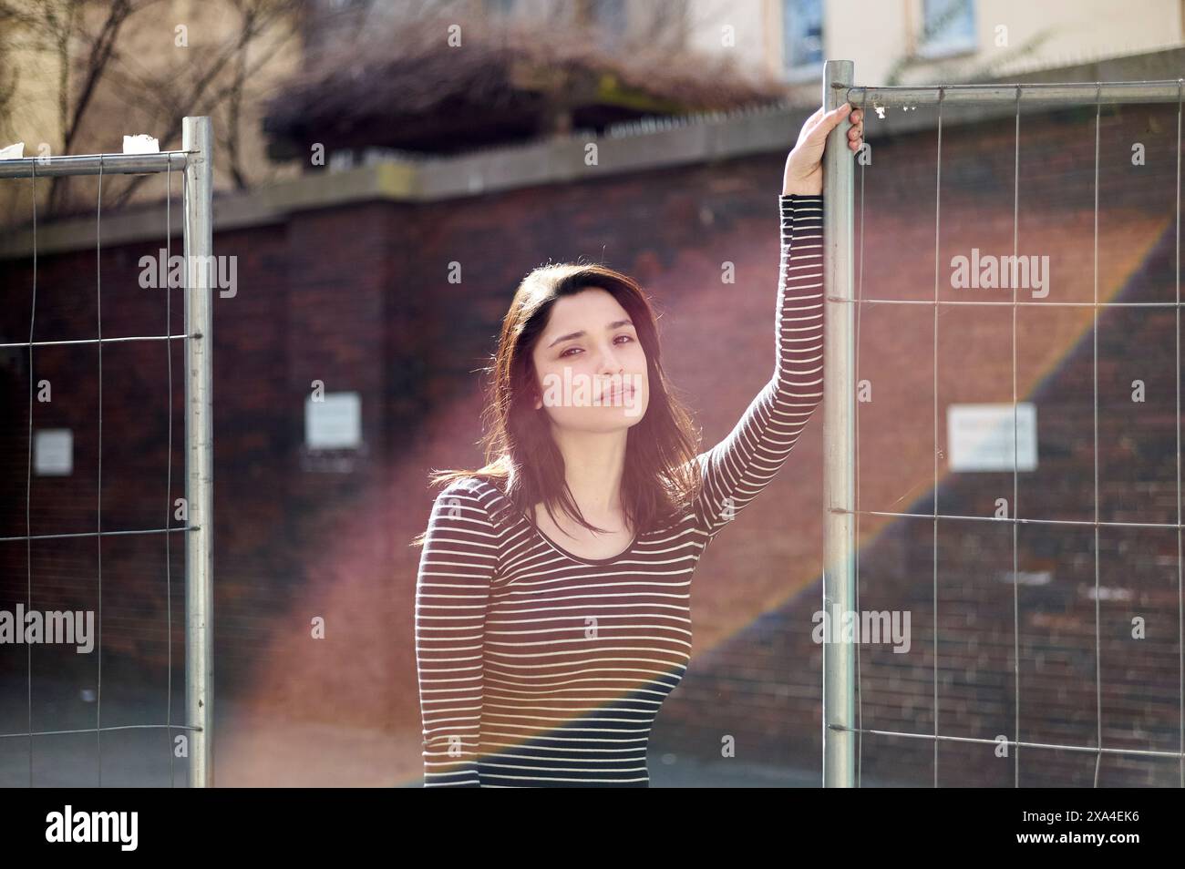 Eine junge Frau in einem gestreiften Oberteil steht an einem Metallzaun und blickt unter natürlichem Sonnenlicht nachdenklich zur Seite. Stockfoto