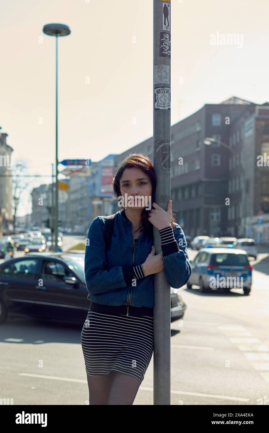 Eine junge Frau steht in sonniger urbaner Umgebung an einer Straßenstange und trägt eine Jeansjacke und einen gestreiften Rock. Stockfoto