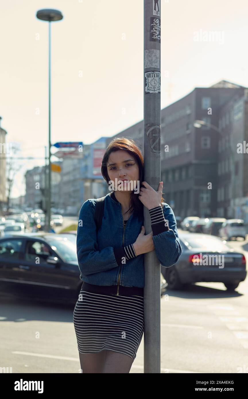 Eine junge Frau lehnt sich an einen Lichtmast auf einer sonnigen Stadtstraße, mit Autos und Gebäuden im Hintergrund. Stockfoto