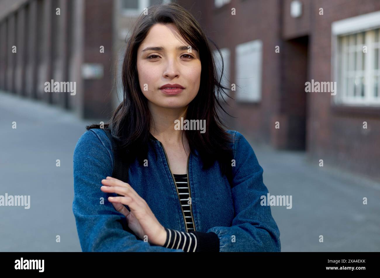 Eine Frau mit dunklen Haaren steht vor einem Ziegelgebäude, trägt eine Jeansjacke mit gefalteten Armen. Stockfoto