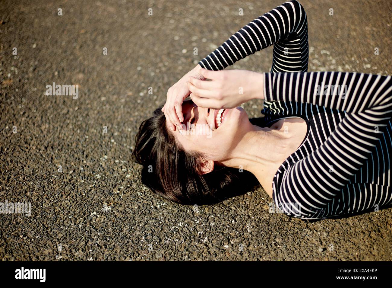 Eine Frau liegt auf dem Rücken auf dem Boden, lacht mit ihren Händen, die teilweise ihr Gesicht bedecken, und trägt ein gestreiftes Top. Stockfoto
