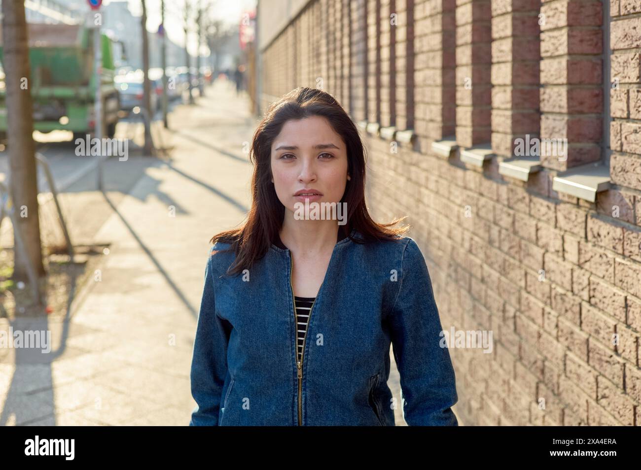 Eine Frau steht auf einem Bürgersteig neben einer Ziegelmauer und blickt bei Tageslicht neutral in die Kamera. Stockfoto