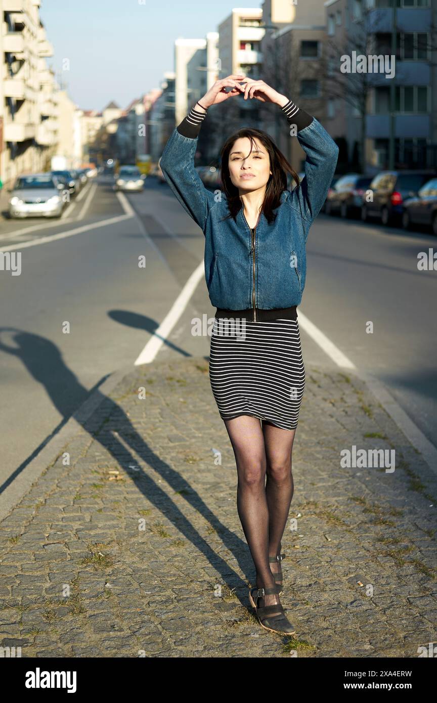 Eine Frau steht tagsüber mitten in einer Stadtstraße und streckt ihre Arme über dem Kopf mit Gebäuden im Hintergrund. Stockfoto