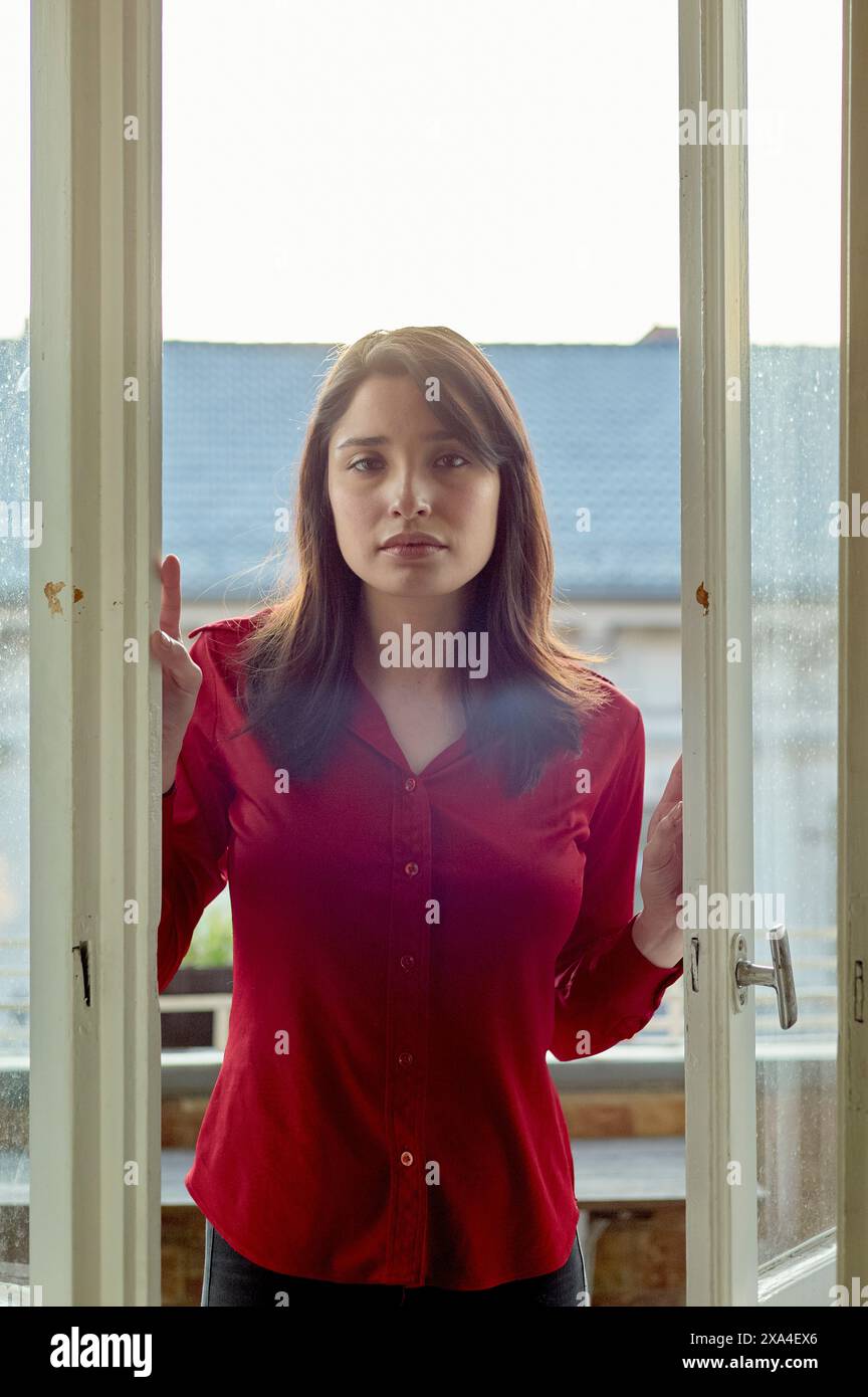 Eine Frau steht zwischen offenen Glastüren, hinterleuchtet mit weichem Licht, das durch transparente Vorhänge filtert, und trägt eine rote Bluse und einen besinnlichen Ausdruck. Stockfoto