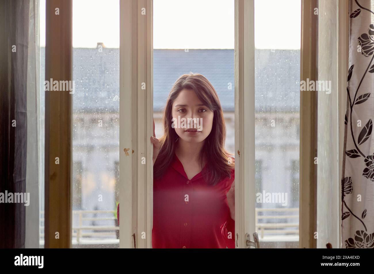 Eine junge Frau steht vor einem Fenster und blickt mit sanftem Blick hinein. Das Fenster ist mit einem gemusterten Vorhang geschmückt, und die Szene hinter der Frau lässt eine städtische Umgebung vermuten. Stockfoto