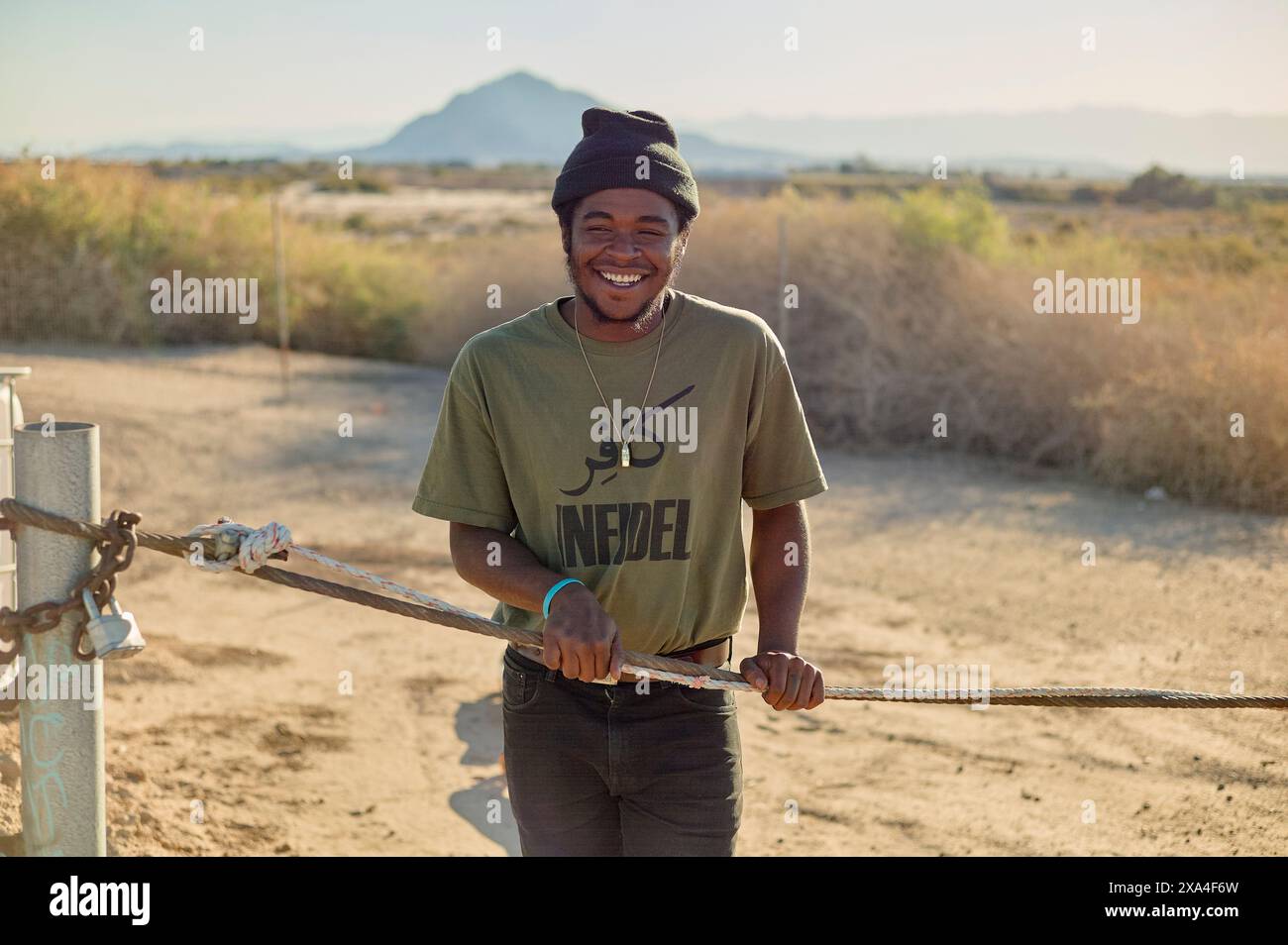 Ein lächelnder Mann mit Mütze und grünem T-Shirt steht hinter einem Metalltor, mit einer sonnendurchfluteten Berglandschaft im Hintergrund. Stockfoto