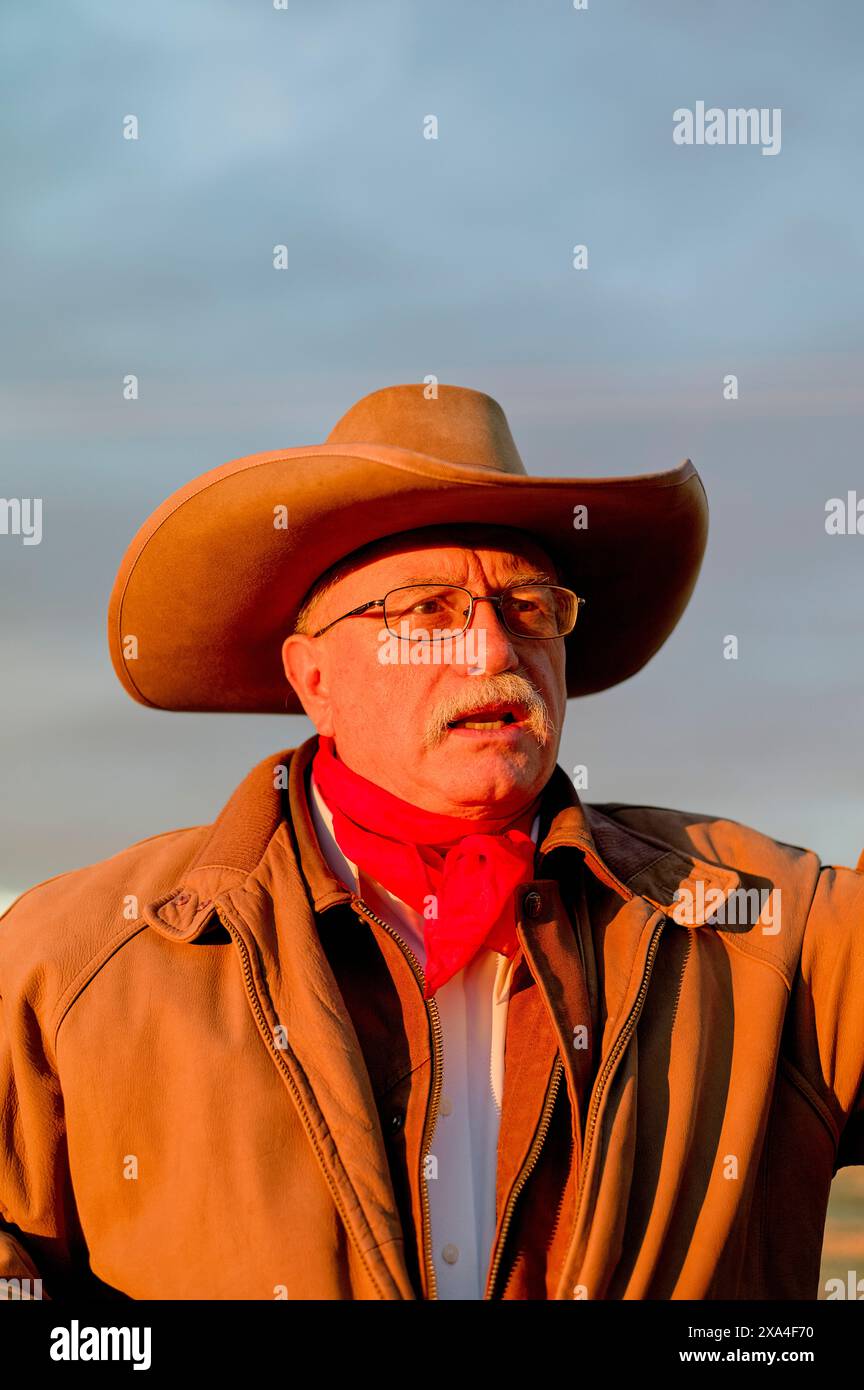 Ein älterer Mann mit Brille und Schnurrbart mit Cowboyhut und rotem Halstuch schaut in die Ferne. Er trägt eine braune Jacke, und der Hintergrund deutet auf Sonnenuntergang hin. Stockfoto