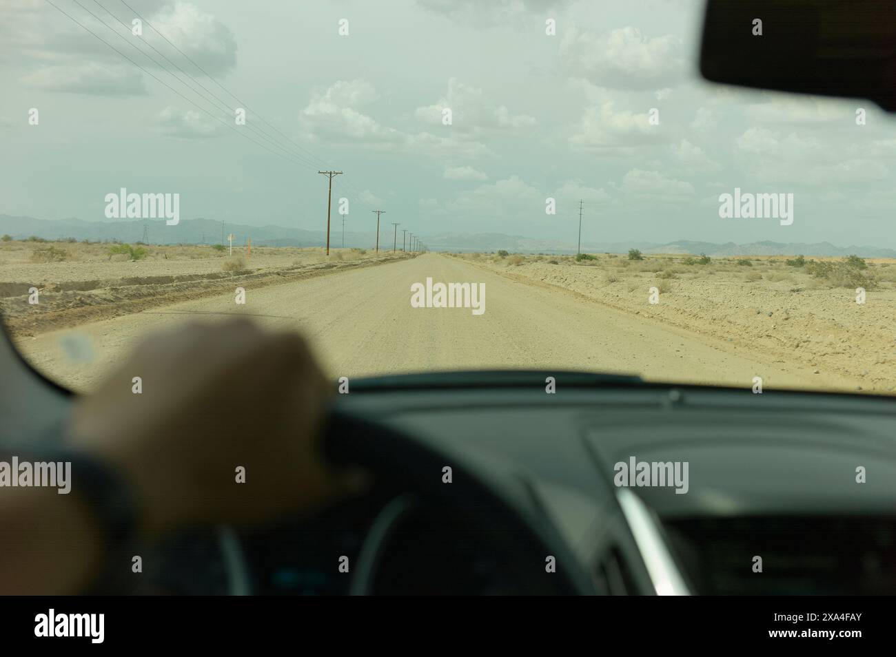 Ein Blick aus dem Inneren eines Fahrzeugs, das eine staubige Wüstenstraße hinunterfährt, mit Telefonmasten, die auf einer Seite unter einem bewölkten Himmel liegen. Stockfoto