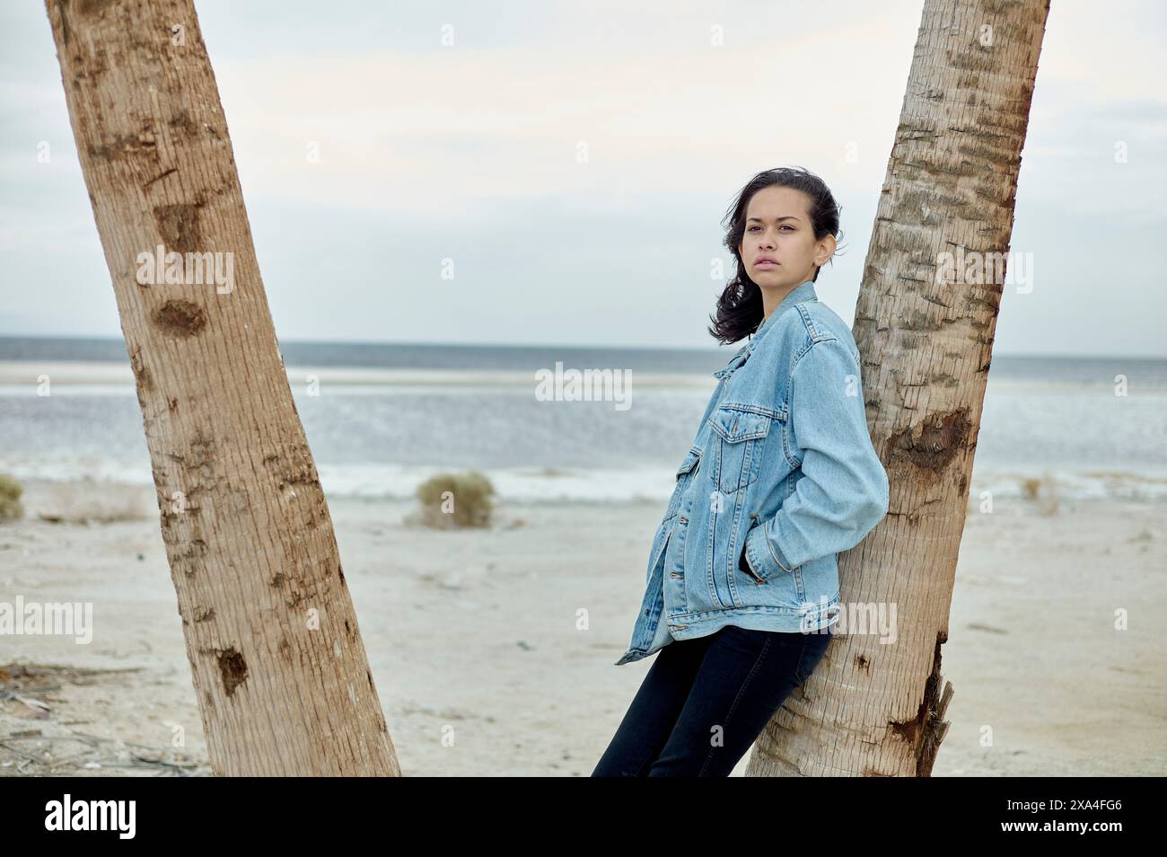 Eine Frau steht an einem bewölkten Tag zwischen zwei Palmen, ihre Arme überkreuzt, während sie mit nachdenklichem Ausdruck in die Ferne blickt. Stockfoto