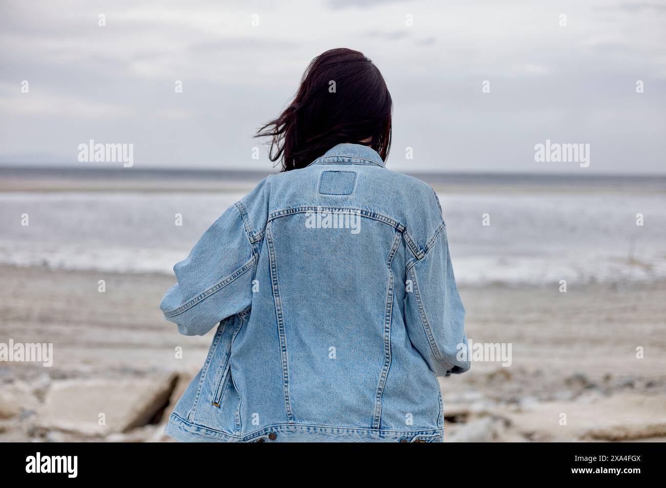Eine Person mit langen dunklen Haaren in einer Jeansjacke steht vor einem ruhigen Meer, mit dem Horizont im Hintergrund unter einem bewölkten Himmel. Stockfoto