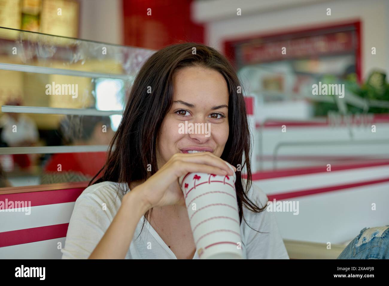 Eine lächelnde Frau ist lässig gekleidet und trinkt aus einem gestreiften Papierbecher mit Deckel in einem Diner mit Retro-Ästhetik. Stockfoto