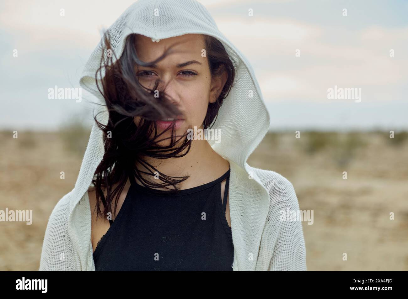 Eine Person mit langen Haaren, die ein weißes Kapuzenkleid trägt, steht vor einer öden Landschaft, mit dem Haar über das Gesicht. Stockfoto