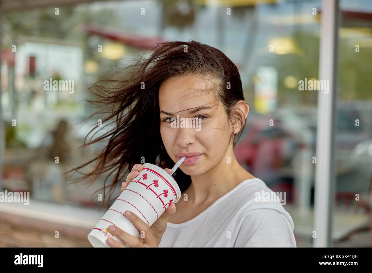 Eine Frau steht draußen und hält einen Drink mit einem Strohhalm an den Mund, ihr Haar weht im Wind, mit einer Ladenfront im Hintergrund. Stockfoto