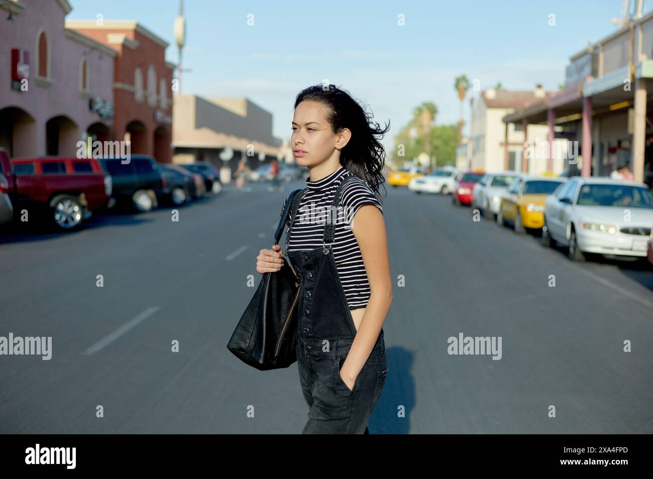 Eine junge Frau läuft über eine Straße in einer sonnigen städtischen Umgebung, trägt eine schwarze Schultertasche und trägt ein gestreiftes Hemd mit Overall. Stockfoto