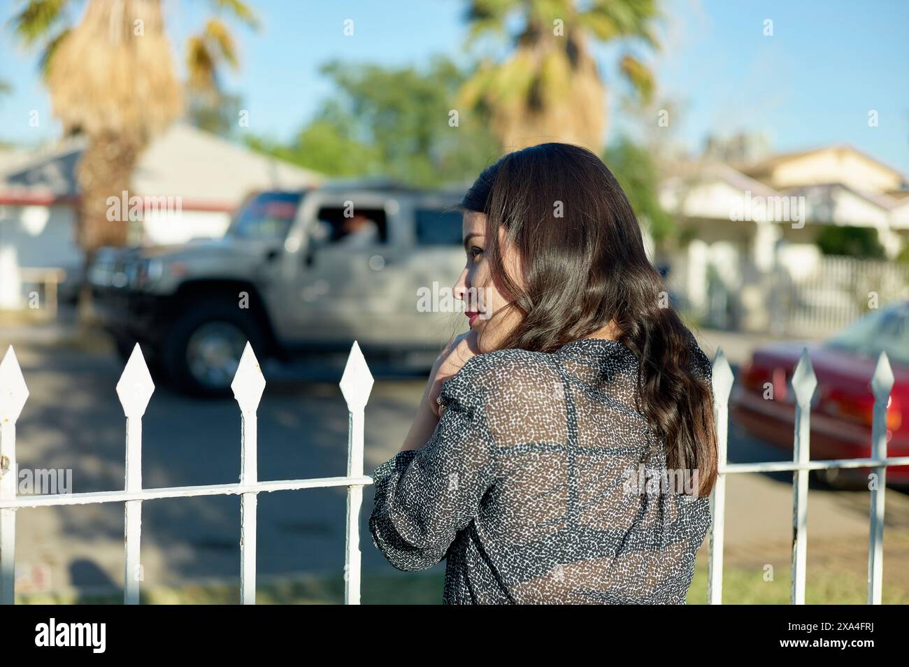 Eine Frau, die an einem weißen Pfostenzaun steht und an einem sonnigen Tag mit Wohnhäusern und Fahrzeugen in die Ferne blickt. Stockfoto