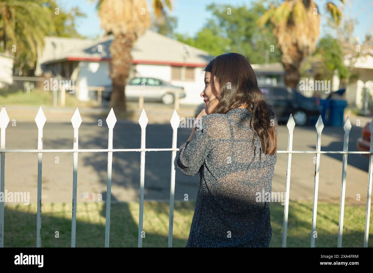 Eine Frau steht hinter einem weißen Pfostenzaun, mit dem Rücken zur Kamera gerichtet, und blickt über ihre Schulter in einem Wohnviertel. Stockfoto