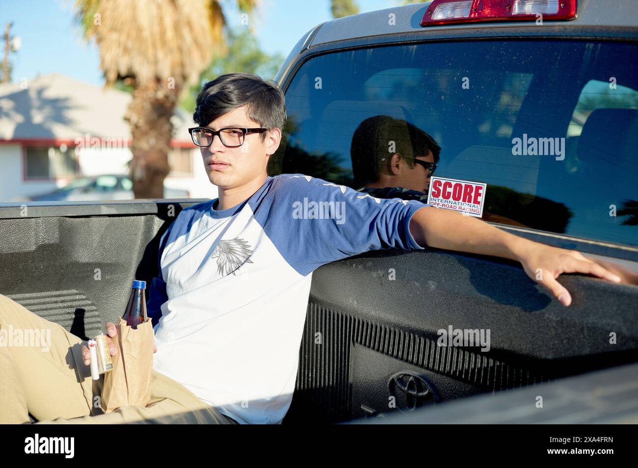 Ein junger Mann mit einer Brille, der ein blau-graues Hemd trägt und eine blaue Flasche hält, sitzt gemütlich auf dem Rücksitz eines Pickup-Trucks, mit einer Wohnstraße und Palmen im Hintergrund. Stockfoto