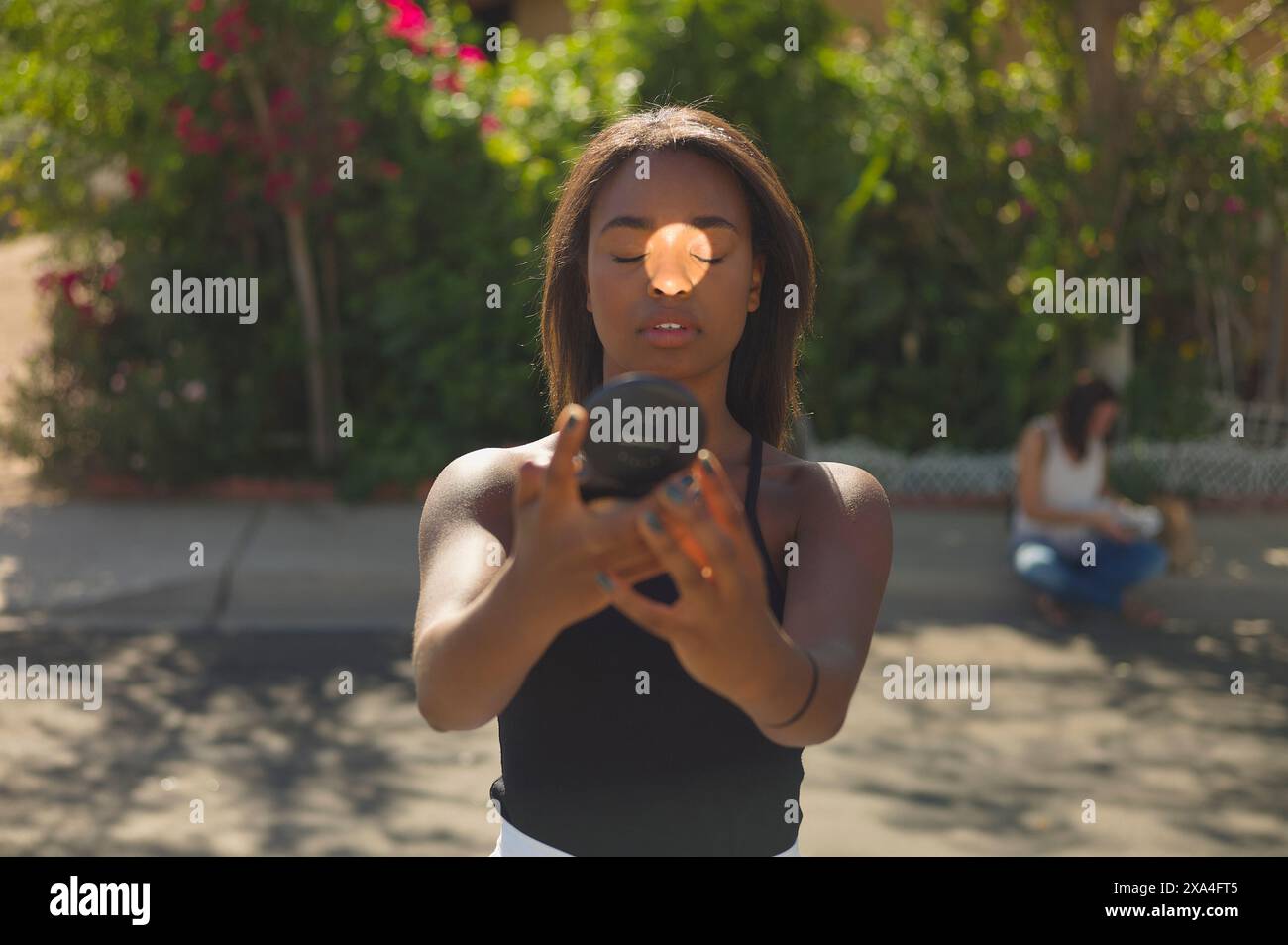 Eine fokussierte junge Frau hält ein Kameraobjektiv an ihr Auge und macht ein Foto, während eine andere Person im Hintergrund auf einer Bank sitzt, umgeben von grünem Laub. Stockfoto