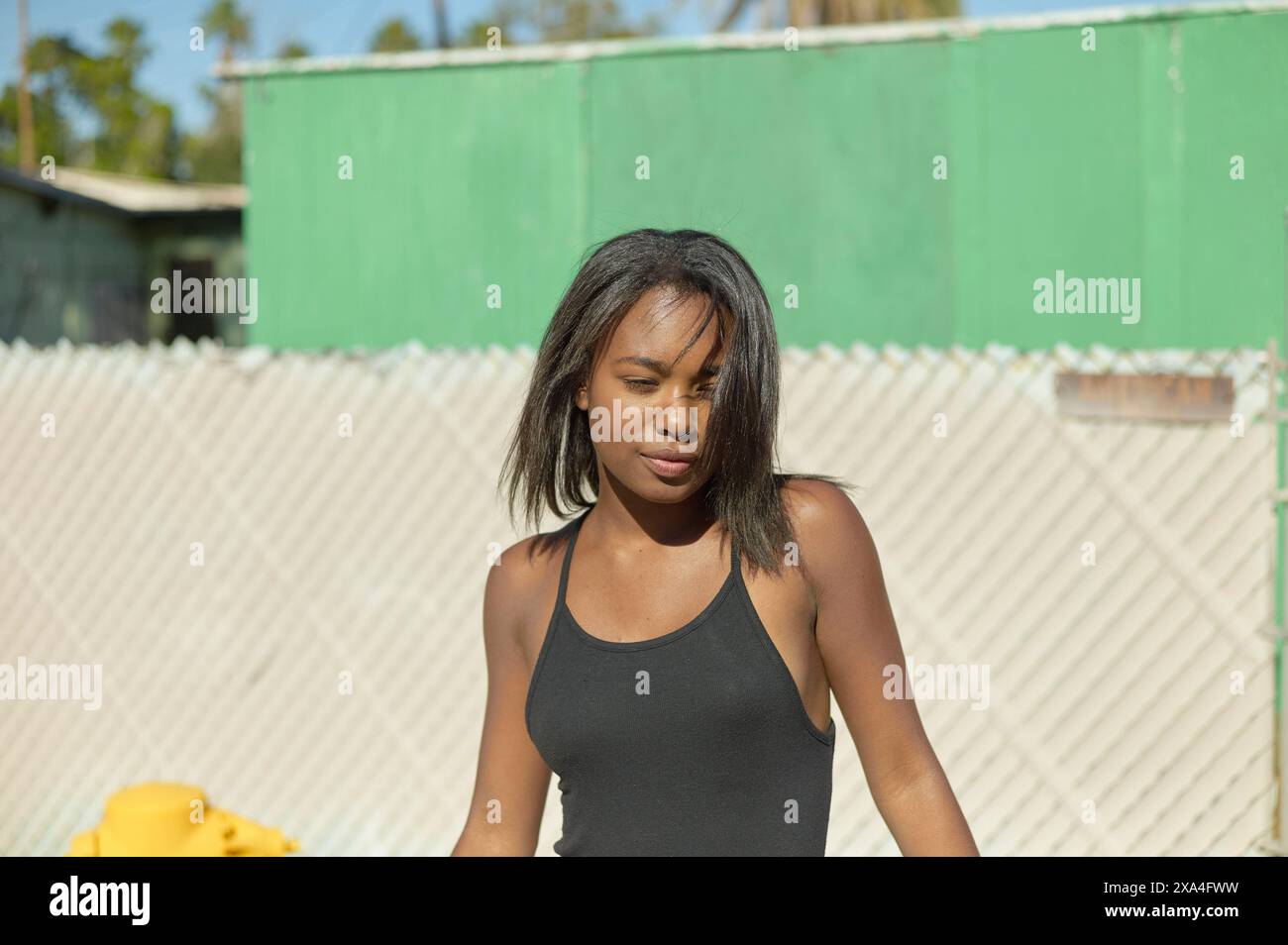 Eine junge Frau mit dunklem Haar, die ein schwarzes Tanktop trägt, steht draußen, schwindelnd im Sonnenschein, mit einem grünen Gebäude und einem Maschendrahtzaun im Hintergrund. Stockfoto