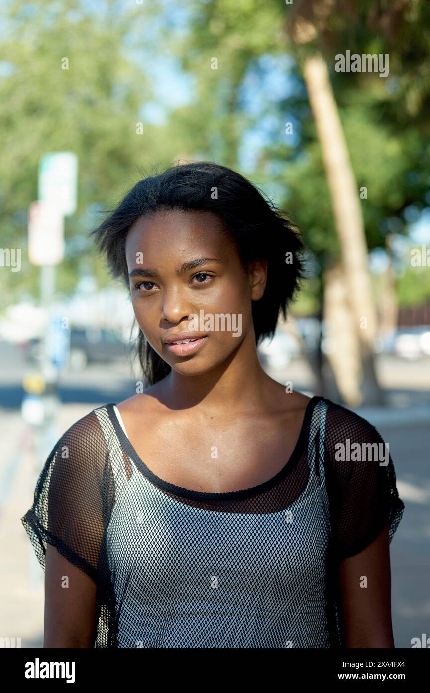 Eine Frau steht draußen an einem sonnigen Tag, mit Bäumen und Straßenschildern im Hintergrund, und blickt entspannt in die Kamera. Stockfoto