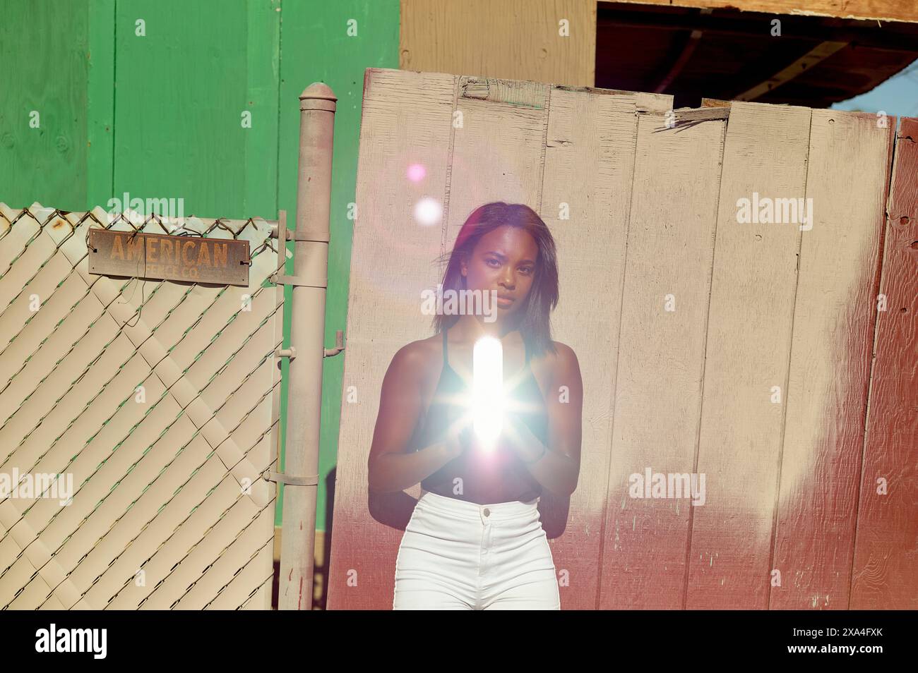 Eine Frau steht vor einer rustikalen Mauer und einem Zaun mit dem Wort „AMERICAN“ auf einem Schild und hält eine helle Lichtquelle, die ihren Mittelteil verdeckt. Stockfoto