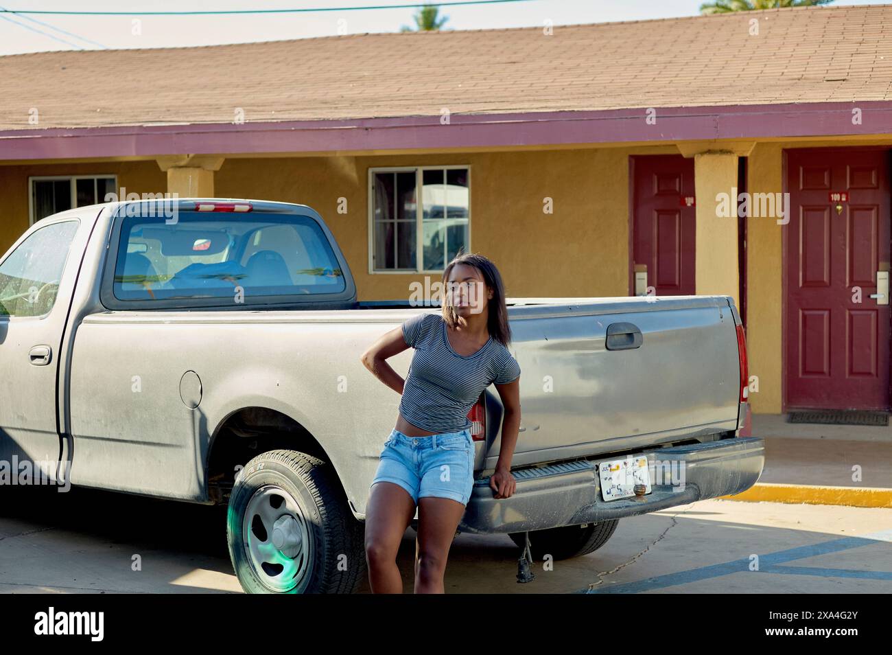 Eine Frau steht vor einem silbernen Pickup-Truck, der bei einem senffarbenen Gebäude geparkt ist, mit einer roten Tür, der Hand auf der Hüfte und einem besinnlichen Gesichtsausdruck. Stockfoto