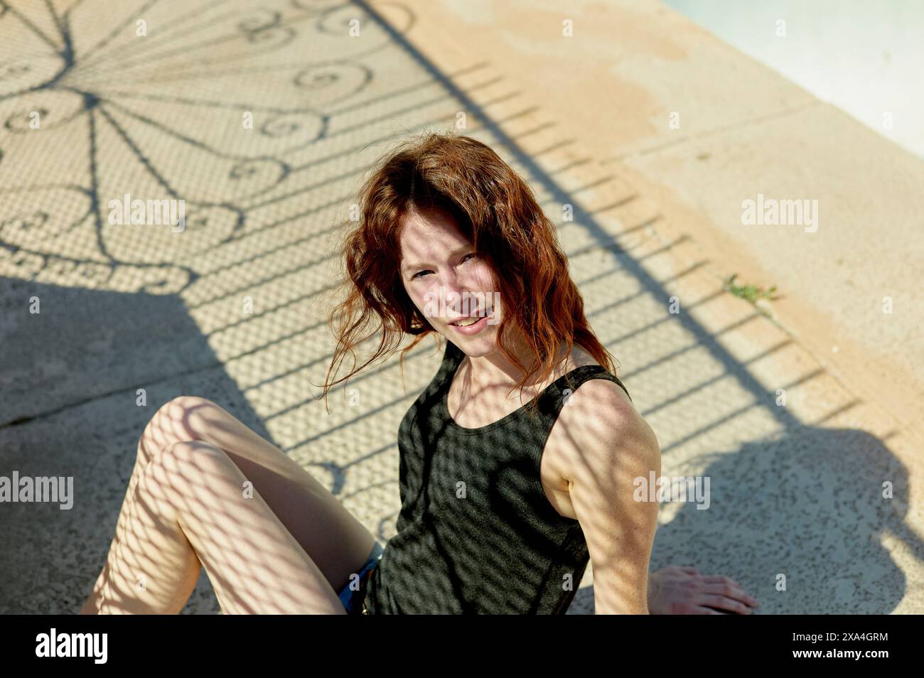Eine Frau mit rotem Haar lächelt, während sie am Pool sitzt, während die Sonne einen Schatten von einem Zaunmuster auf sie und den Boden wirft. Stockfoto