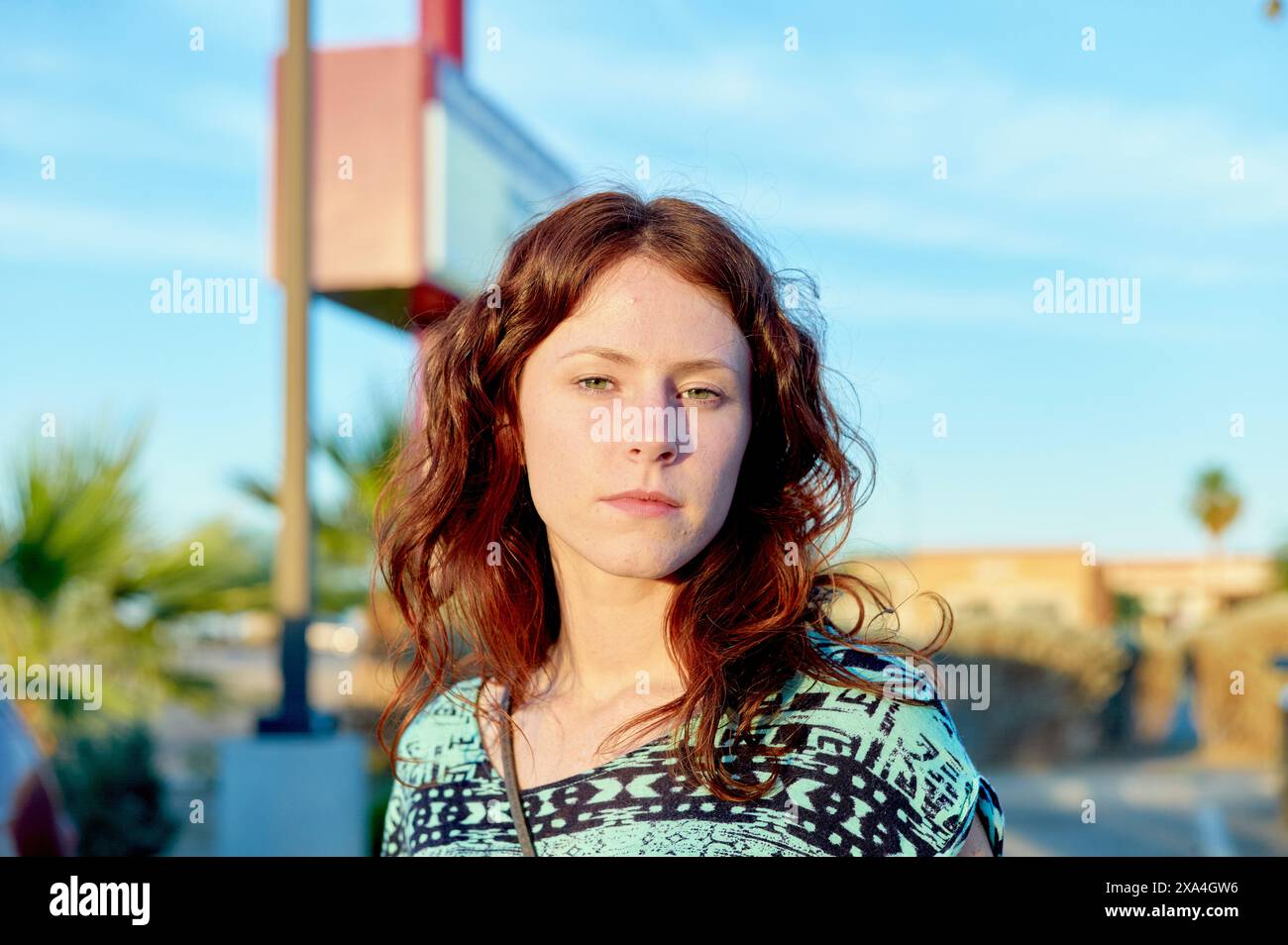 Eine Frau mit rötlich-braunem Haar steht draußen, ihr Ausdruck nachdenklich, während sie leicht nach links blickt, der Sonnenschein ein warmes Licht auf ihr Gesicht wirft. Stockfoto