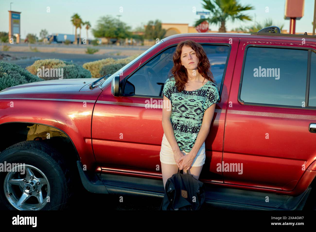 Eine Frau lehnt sich an einem sonnigen Tag mit klarem blauem Himmel an einen roten Pickup-Truck und landschaftlich begrüntem Grün im Hintergrund. Stockfoto
