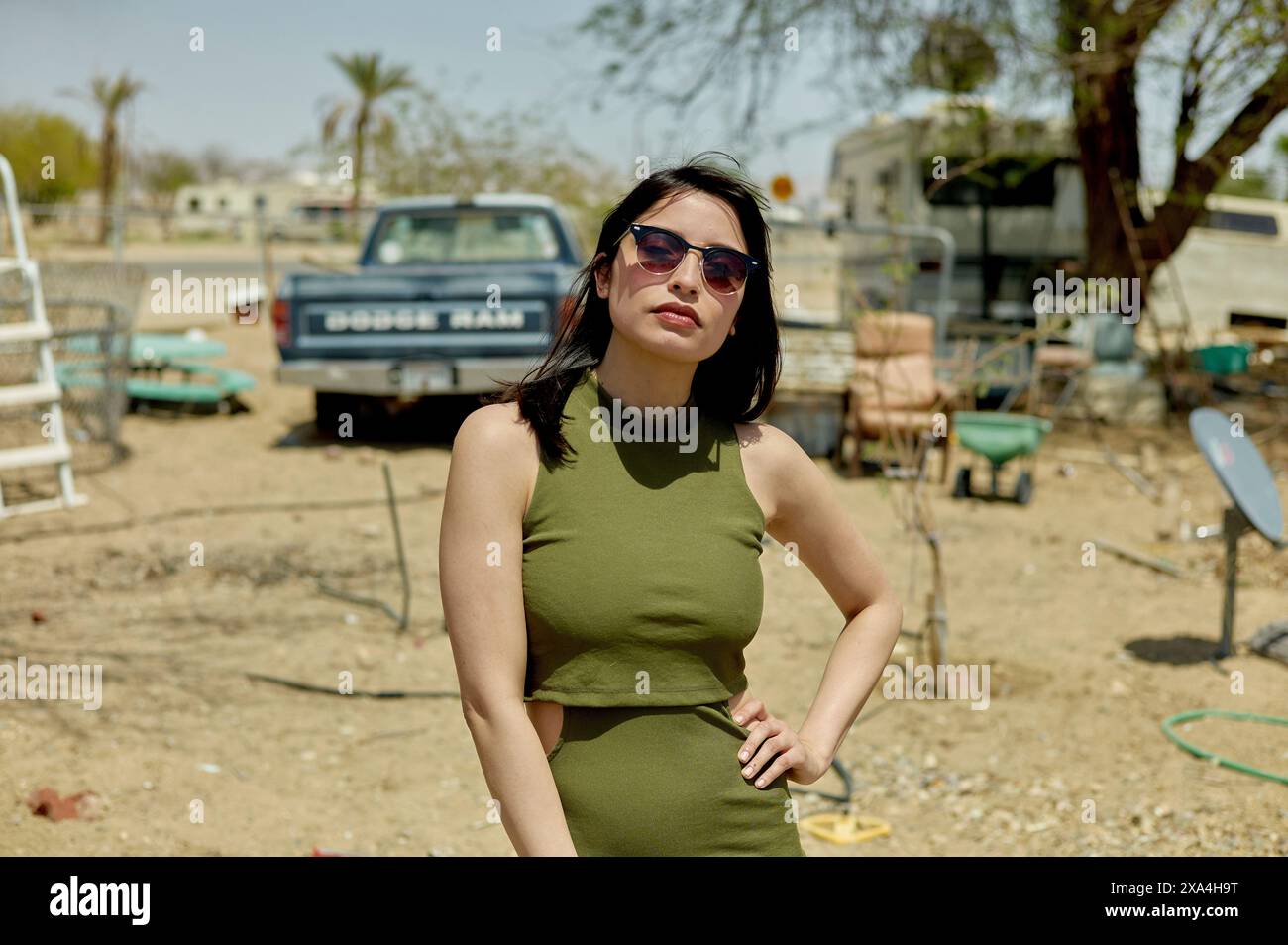 Eine Frau steht draußen in einer sonnigen Gegend mit Anhängern und einem Fahrzeug im Hintergrund. Sie trägt ein ärmelloses Kleid und eine Sonnenbrille. Stockfoto