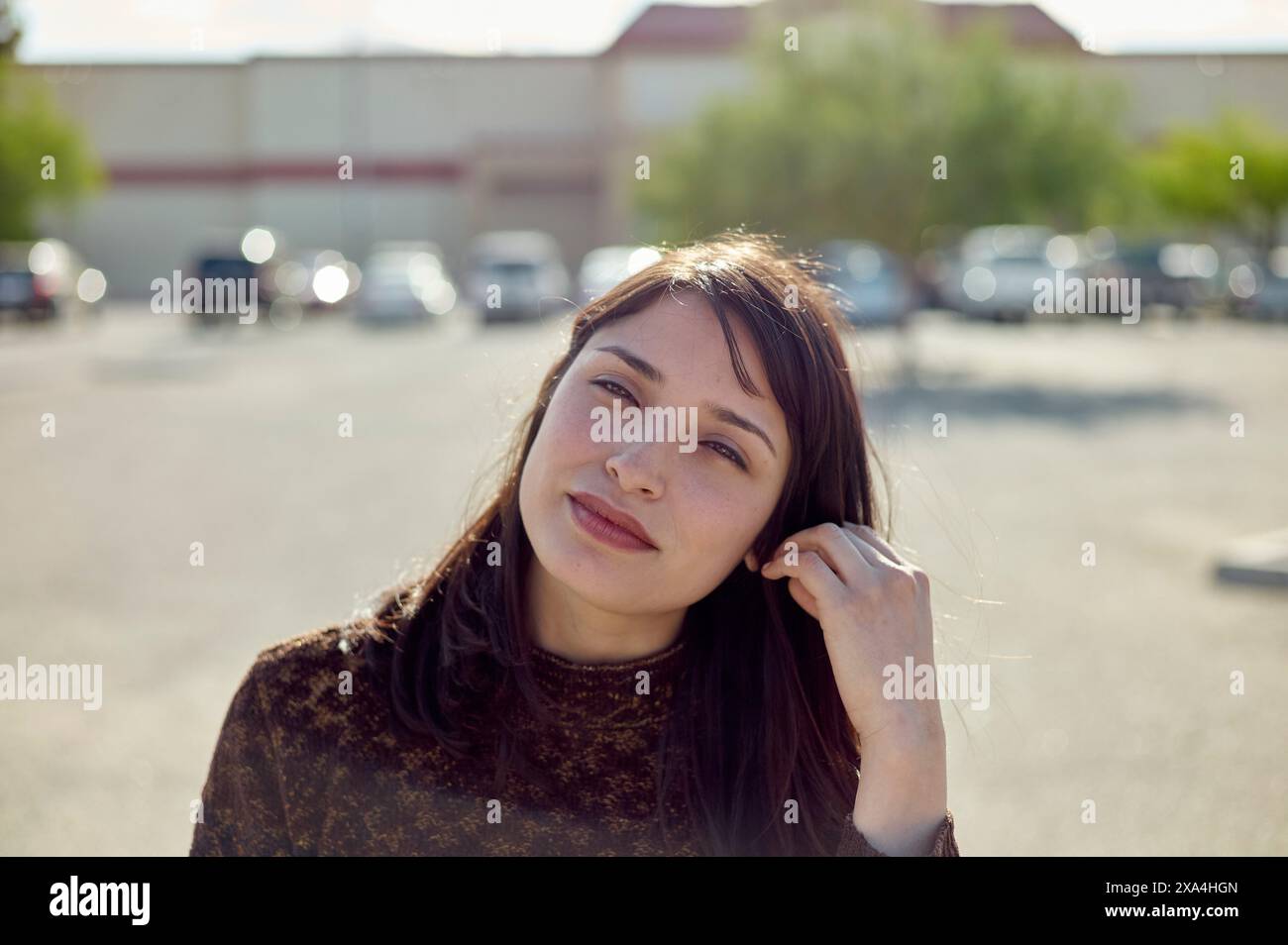 Eine junge Frau mit langen braunen Haaren, die an einem sonnigen Tag auf einem Parkplatz dezent in die Kamera lächelt. Stockfoto