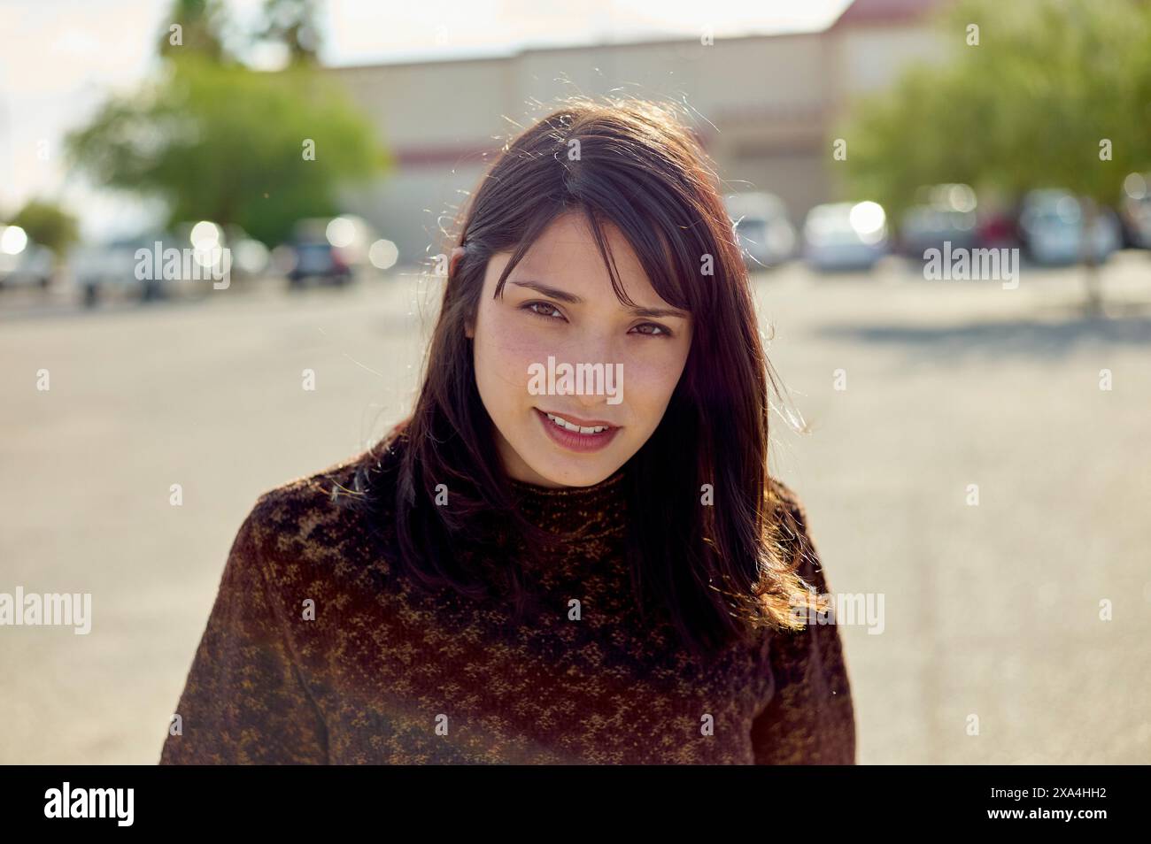 Eine lächelnde Frau mit dunklem Haar, die ein Spitzentop trägt, steht draußen an einem sonnigen Tag, mit Sonnenlicht, das ihr Haar hervorhebt. Stockfoto