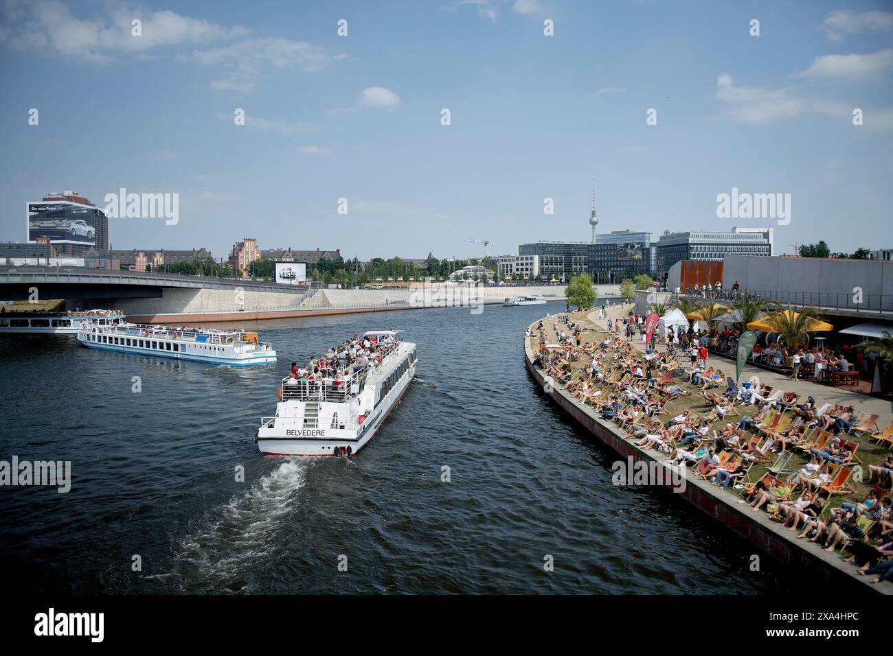 Eine belebte Flusslandschaft mit Menschen, die das sonnige Wetter auf Liegestühlen genießen, mit einem Ausflugsboot, das vorbeisegelt, und einer Stadtlandschaft im Hintergrund. Stockfoto