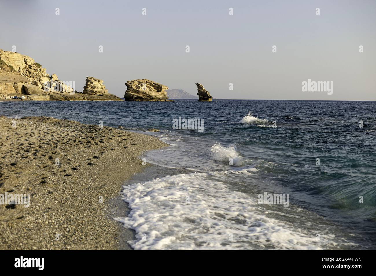 Ruhiger Strand mit sanften Wellen, die sich auf die Sandküste stürzen, mit zerklüfteten Klippen im Hintergrund unter klarem Himmel. Stockfoto
